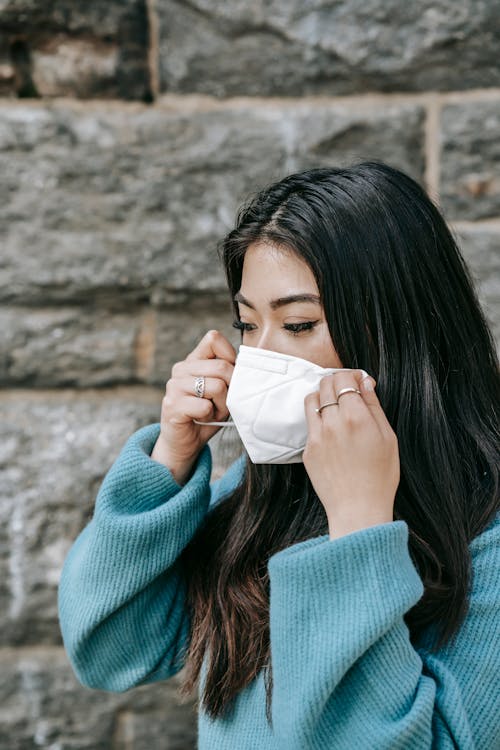 Young Asian female with dark long hair in warm sweater putting white protective mask on face