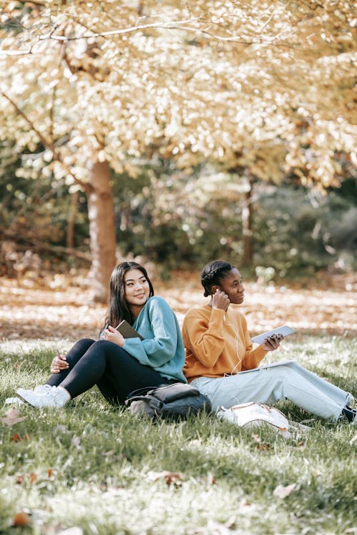 Free Smiling young diverse female students sitting on grassy ground in park and using gadgets while working in home task together and looking away Stock Photo