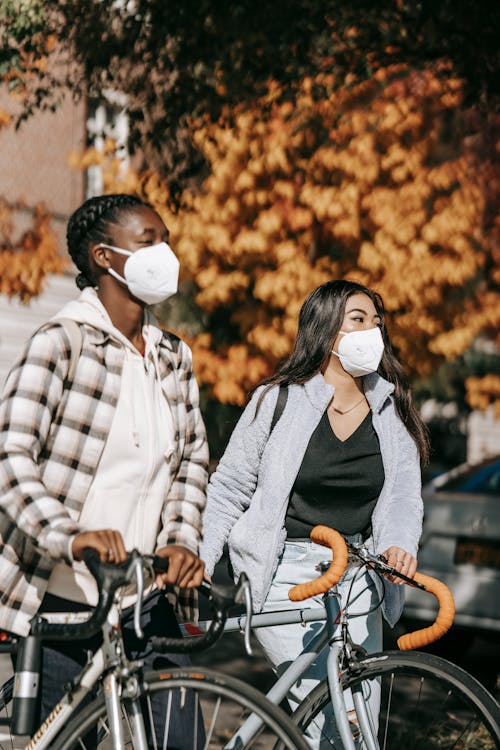 Young multiracial female friends in casual wear and white protective masks with backpacks walking near trees and car with bicycles