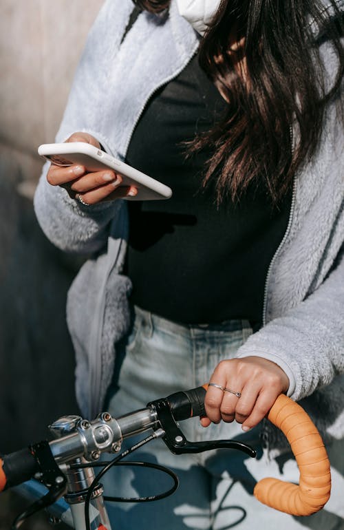 Crop woman in mask text messaging on smartphone standing with bike