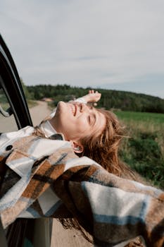 Joyful young woman leans out of car window, embracing freedom during a road trip.