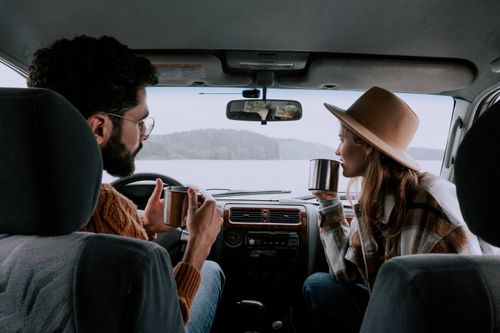 Free Couple enjoying hot drinks inside car with scenic lake view. Perfect for travel and lifestyle themes. Stock Photo