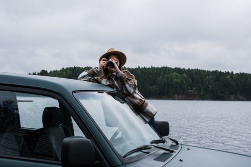 Free Woman in a checkered shirt and hat photographing a scenic lake view from a car. Stock Photo