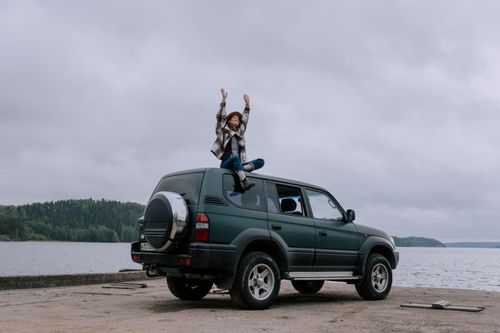 Free A woman sits on top of an SUV with arms raised in excitement by a scenic lake. Stock Photo