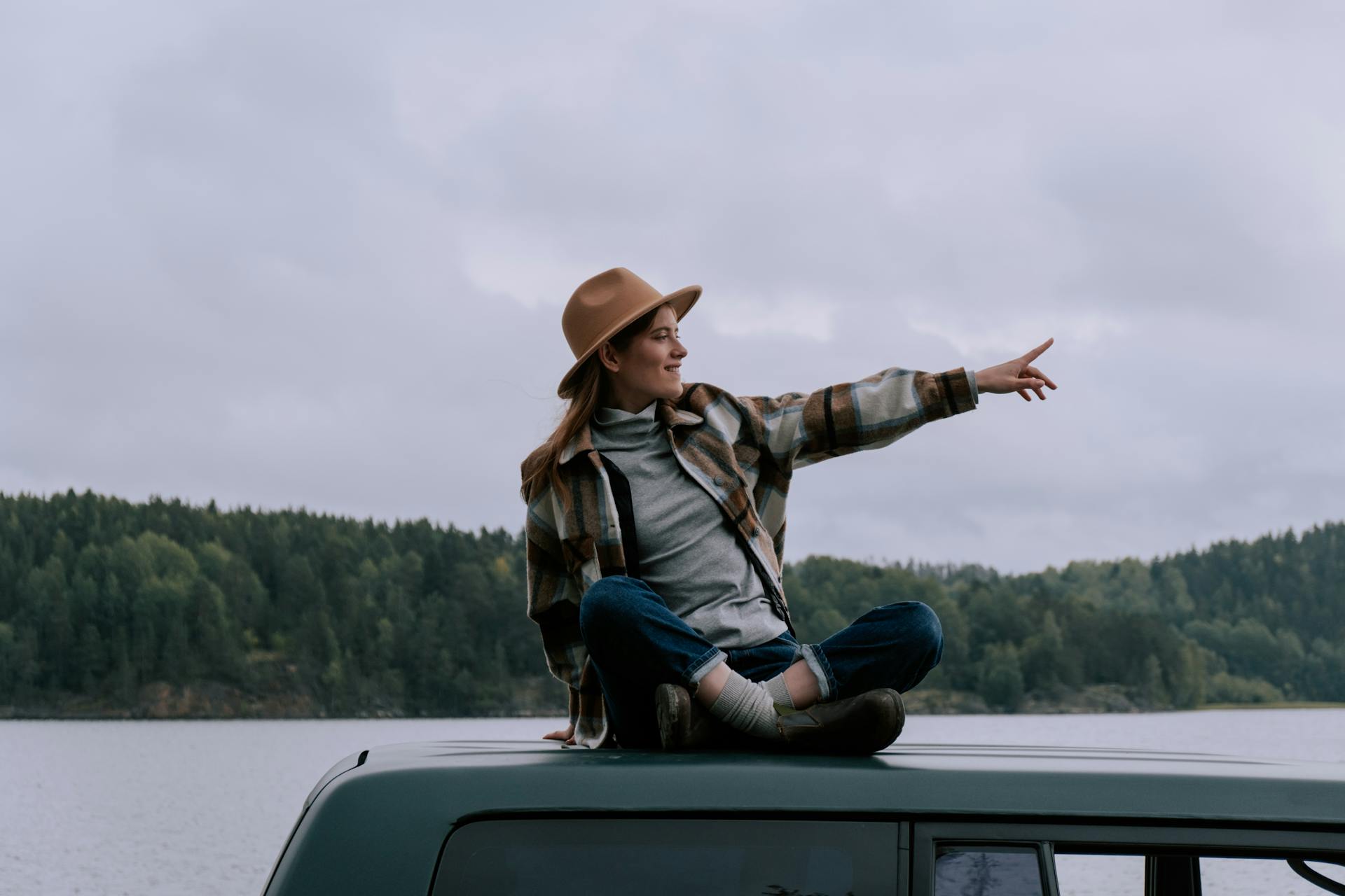 A young woman sitting on a vehicle roof by a lake, pointing and smiling against a forest backdrop.