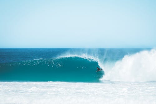 Distant surfer riding on foamy wave in azure ocean against cloudless sky while training in tropical resort on summer day