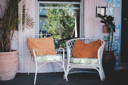 Comfortable wicker chairs with pillows placed on rural house terrace near window and potted houseplants on sunny day