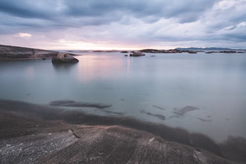 Magnificent scenery of tranquil rocky shore near tranquil seawater reflecting cloudy sky in overcast evening