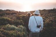 Back view unrecognizable friends wearing casual clothes strolling through green verdant valley covered with thick shrubs towards calm sea in early evening