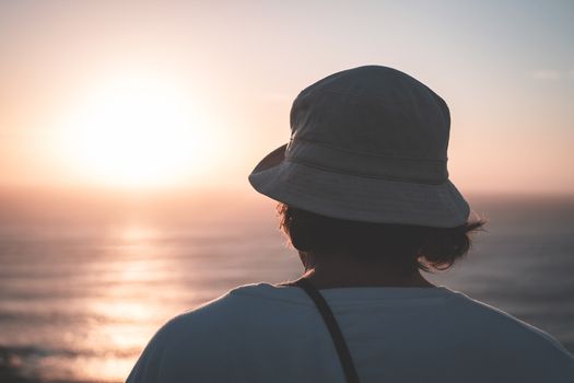 一个戴着夏帽、面目全非的男子站在海岸上，欣赏着日落时分天空下波光粼粼的大海的壮丽景色