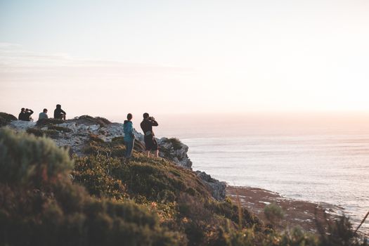 Full length travelers wearing casual clothes resting on rocky cliff and enjoying amazing scenery of calm rippling sea during amazing sunset