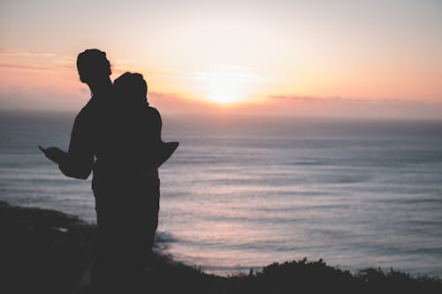 Back view of silhouette of anonymous couple embracing each other while standing on seashore at sundown