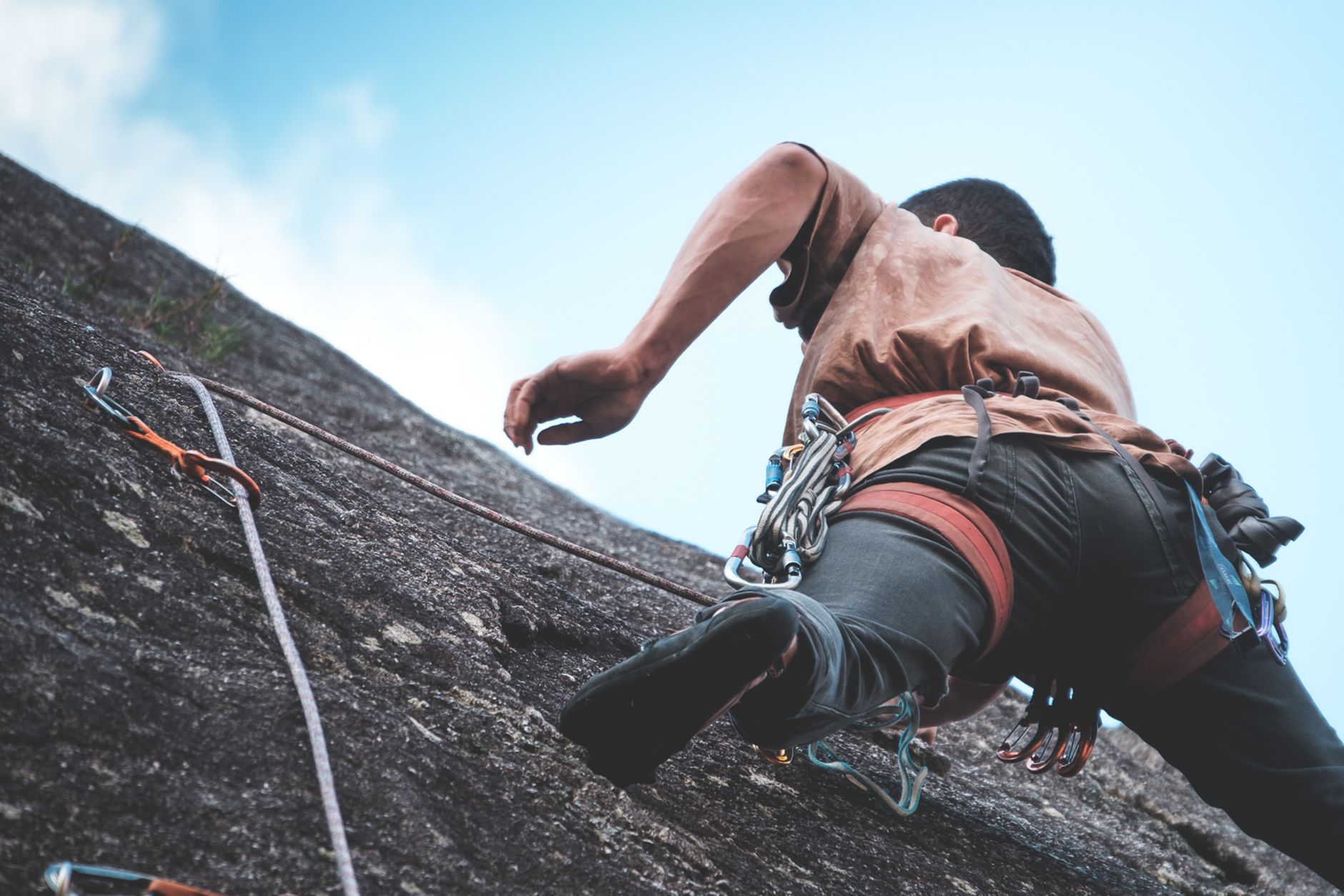 From below back view of anonymous climber in safety harness and special equipment climbing rocky slope against blue sky in mountainous area