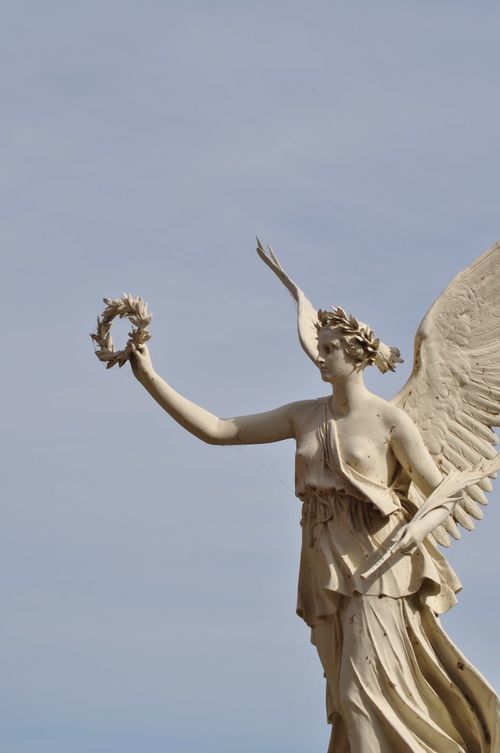 Free An elegant baroque-style angel statue holding a laurel wreath, captured outdoors with a clear sky backdrop. Stock Photo