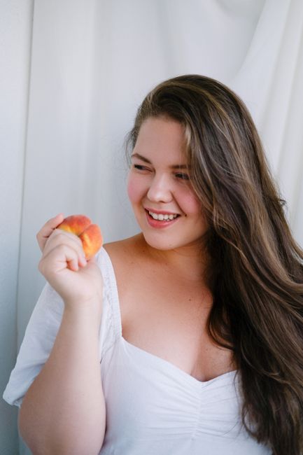 Cheerful plus size female wearing dress smiling and looking away while standing near white curtain with bent arm and fruit in hand