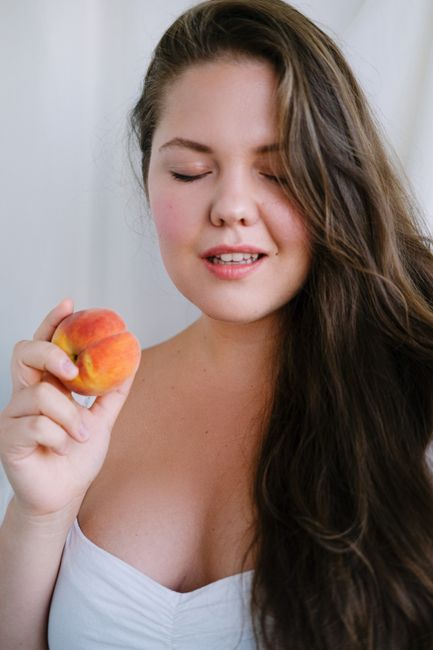 Dreamy female with long hair standing on white background with closed eyes and arm bent in elbow with fresh fruit in hand