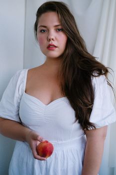 一位冷静而丰满的黑发女性，身着白色连衣裙，站在白色窗帘旁，注视着镜头