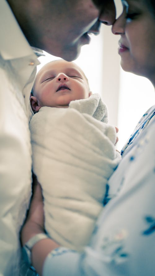 Side view of crop mother in casual clothes standing with newborn sleeping baby and touching noses with husband in daytime
