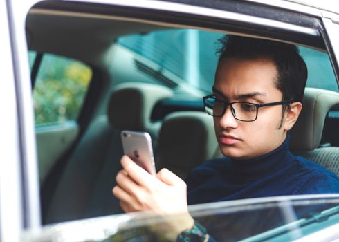 Young man with glasses using smartphone inside a parked car.