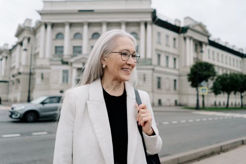 Free A senior woman in a white blazer smiling while walking outdoors near a historic building. Stock Photo