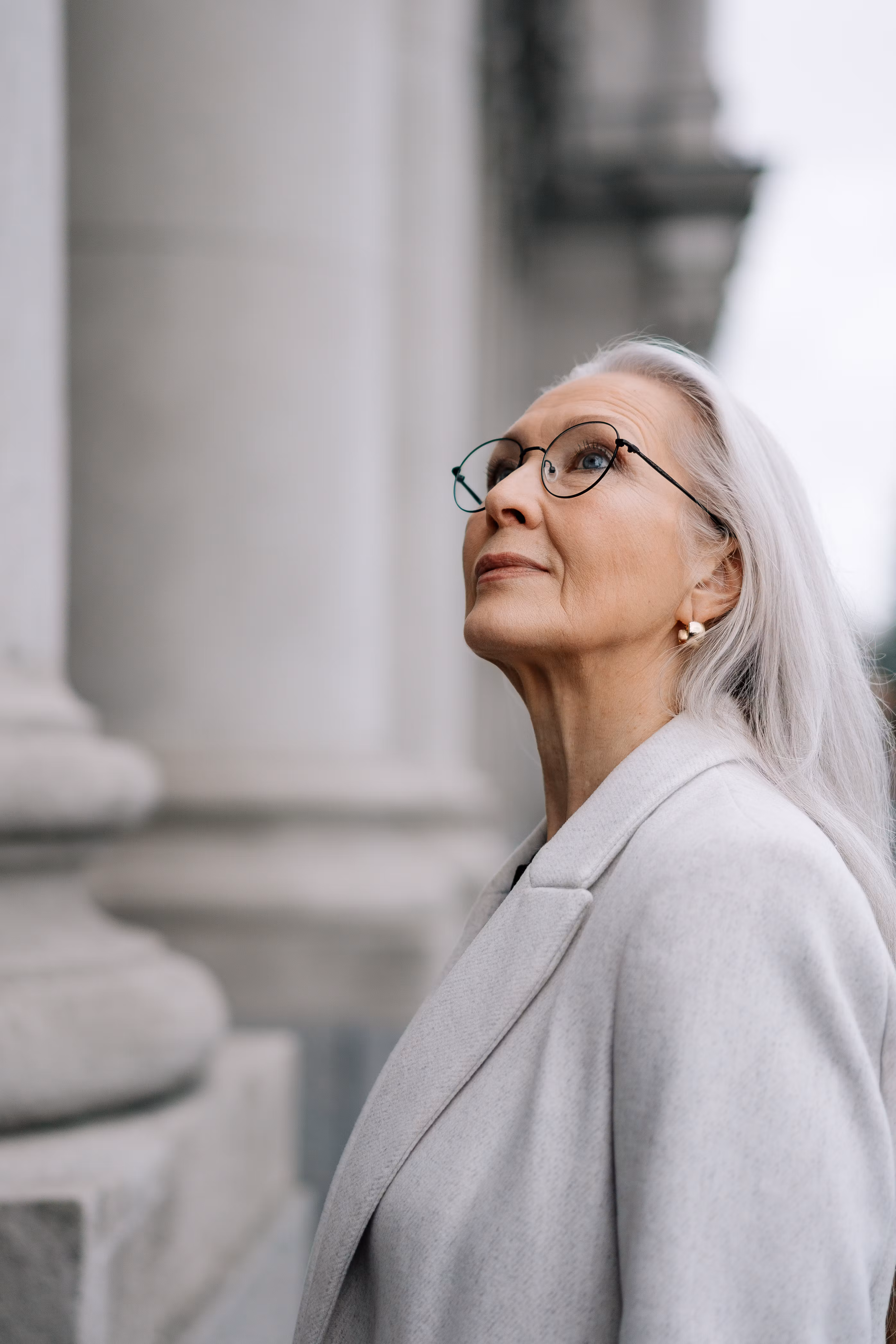 一位戴着眼镜、气质优雅的年长白人妇女，若有所思地抬头望着一栋历史建筑