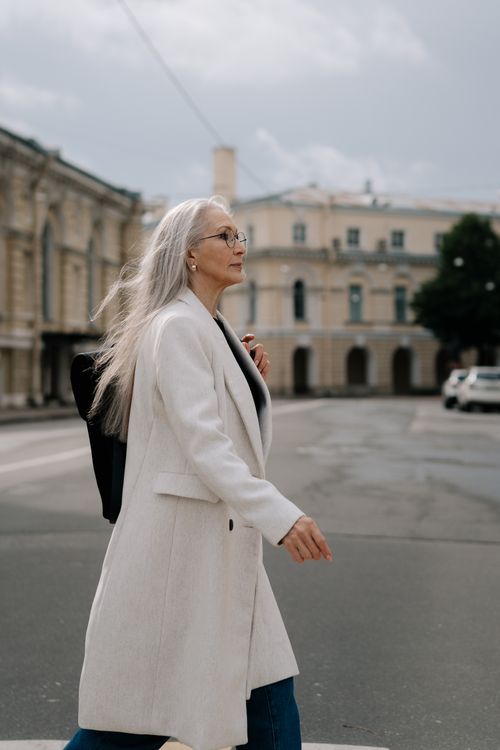 Free Elegant elderly woman with long gray hair walking in a city street, wearing a white coat. Stock Photo