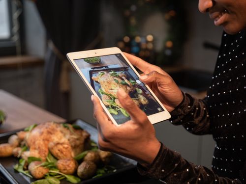 Free Person photographing a roasted chicken and potatoes with a tablet, highlighting food photography. Stock Photo
