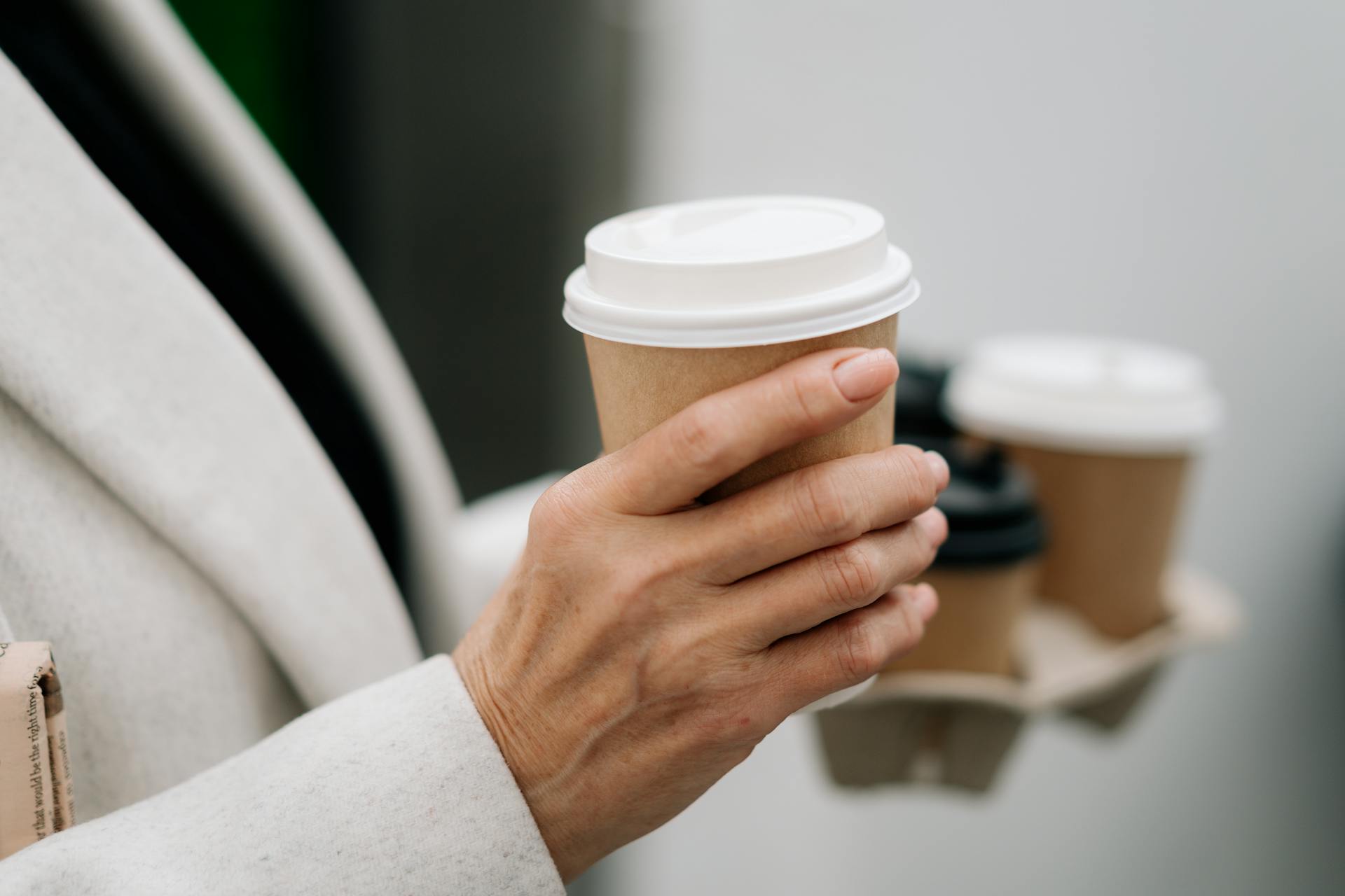 Person holding a cardboard takeaway coffee cup indoors, with two additional cups in a carrier.