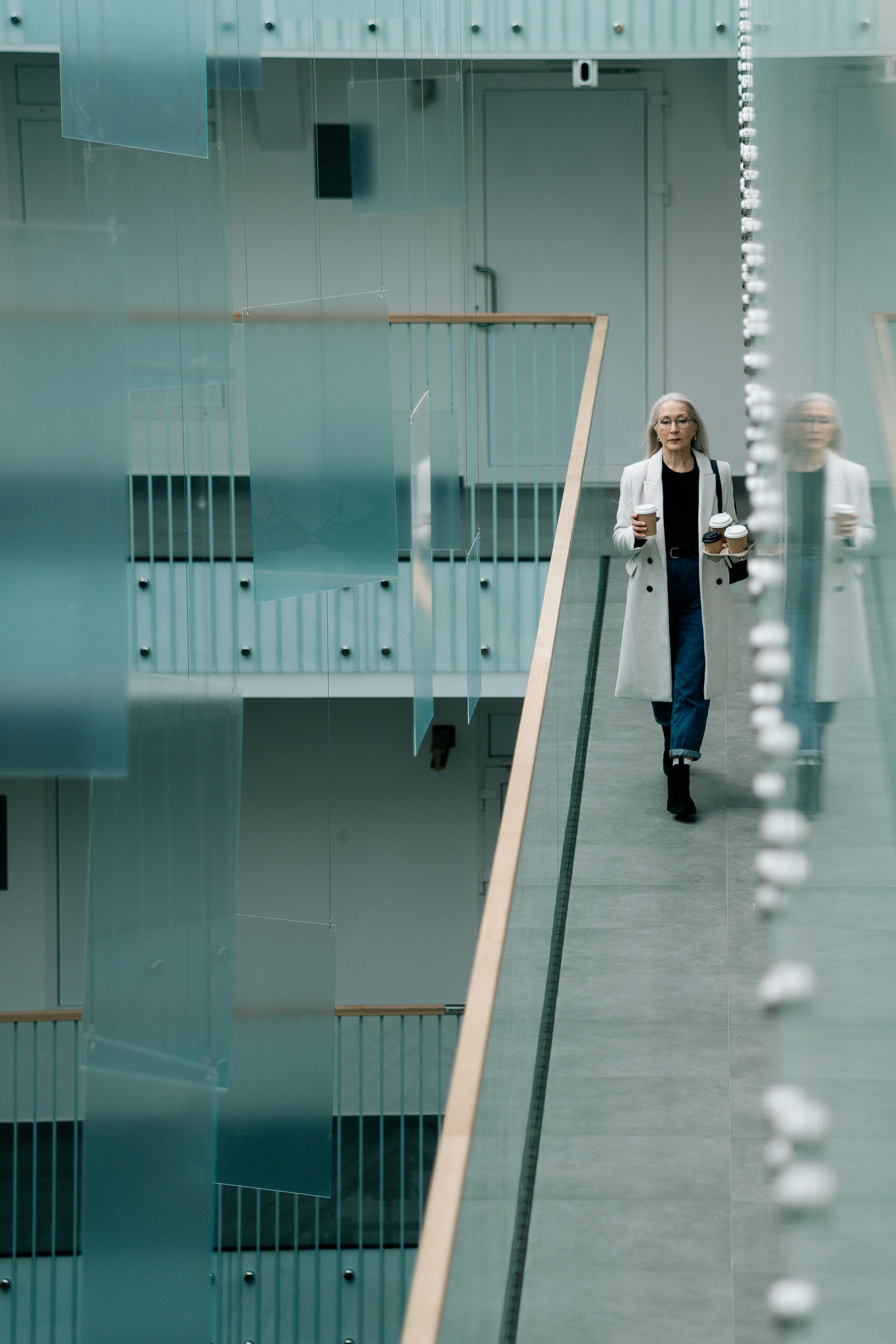 Woman walking in a sleek indoor office corridor with coffee and laptop, embodying professional lifestyle.