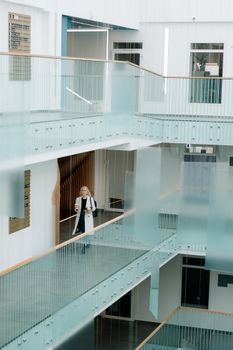 一位女士端着咖啡和文件，走过明亮的现代办公室走廊，走廊装有玻璃栏杆