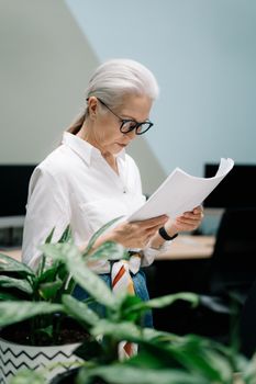 在现代办公室里，一位戴眼镜的老妇人正评论着论文，周围环绕着植物