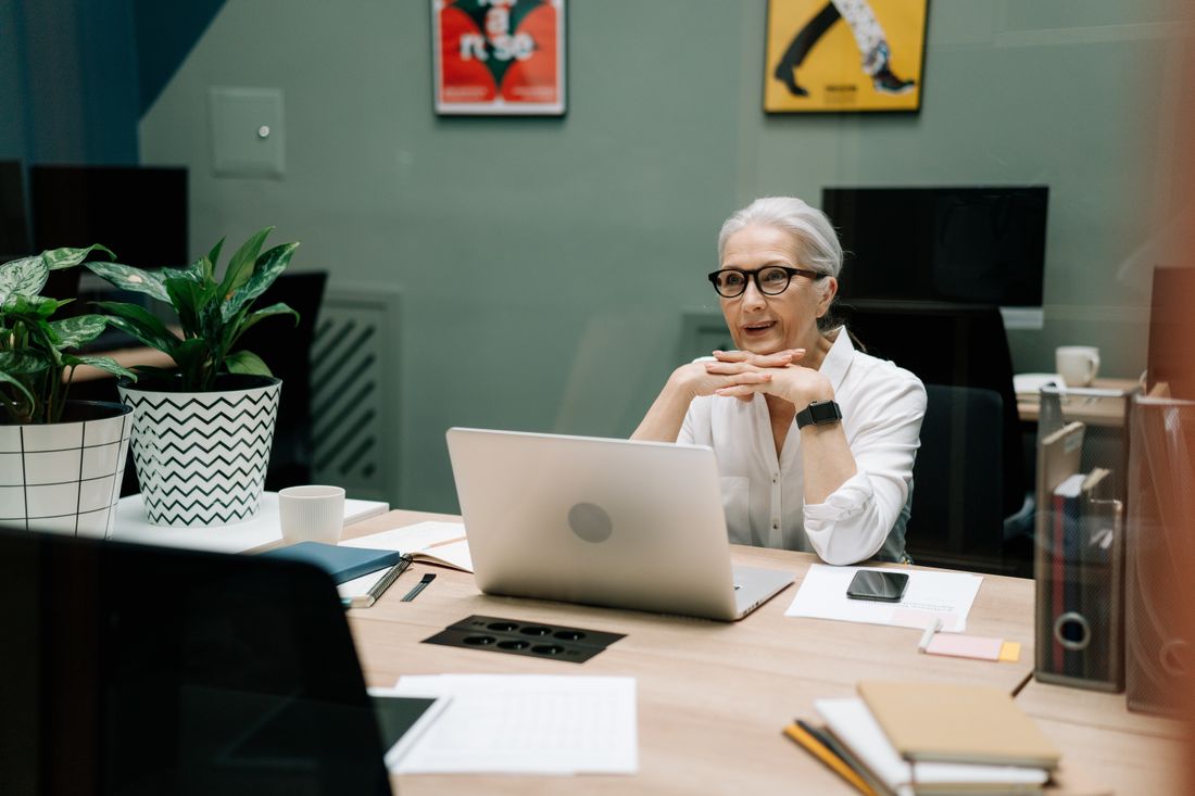 An elderly businesswoman in an office setting, smiling while using a laptop