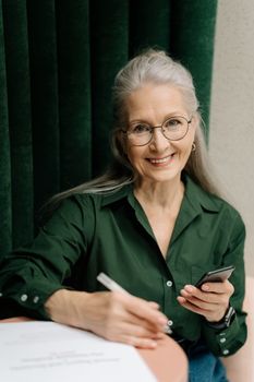一位灰发戴眼镜的优雅女士在室内拿着智能手机微笑