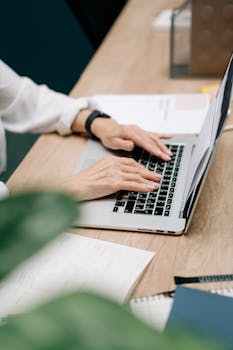 An adult typing on a laptop at a desk, in a well-lit office space with papers and plants nearby.