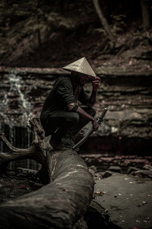 Free A man with a conical hat kneeling on a log with a sword in a forest. Stock Photo
