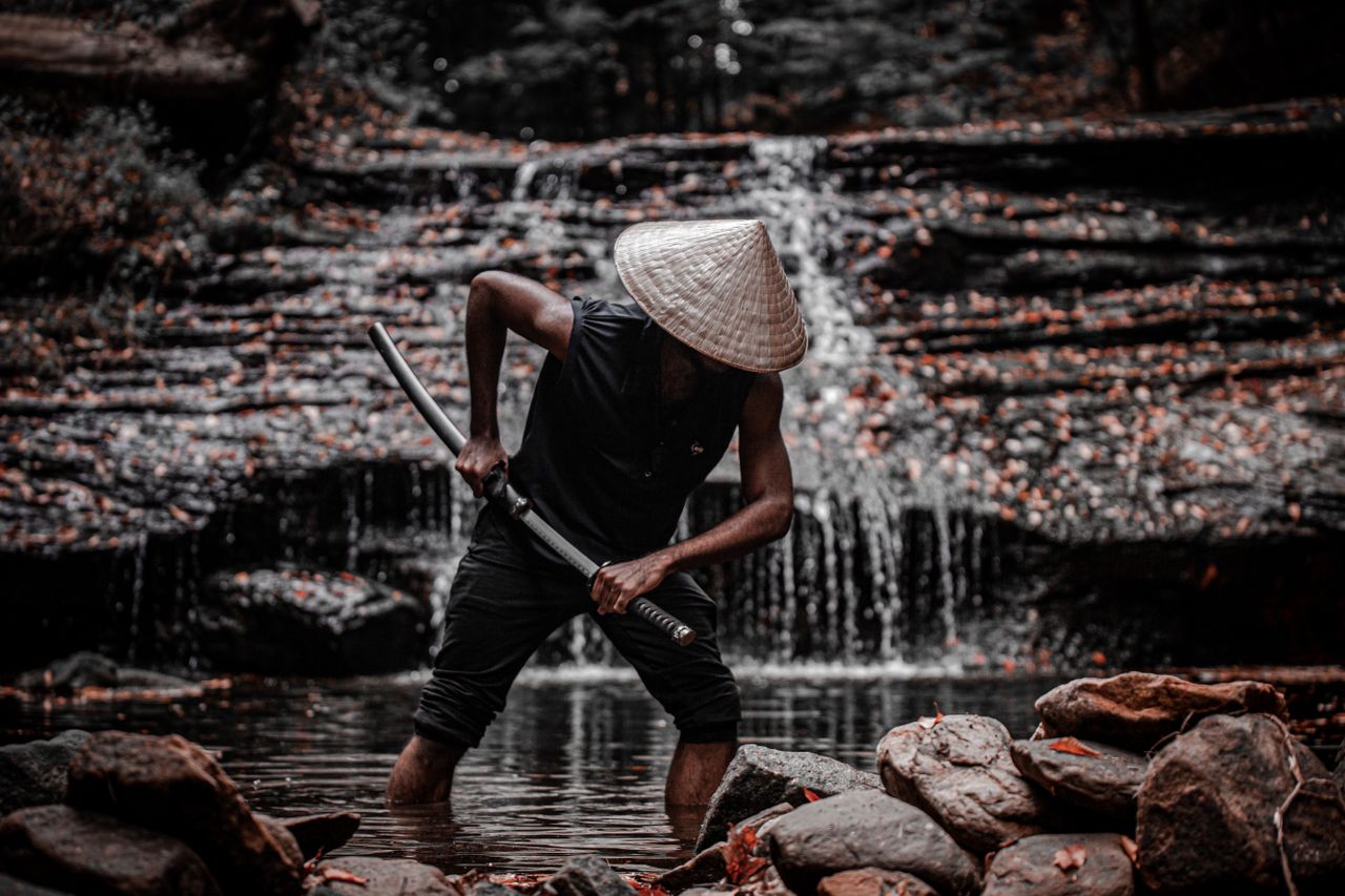 a person in a straw hat with a sword stands in a tranquil waterfall se