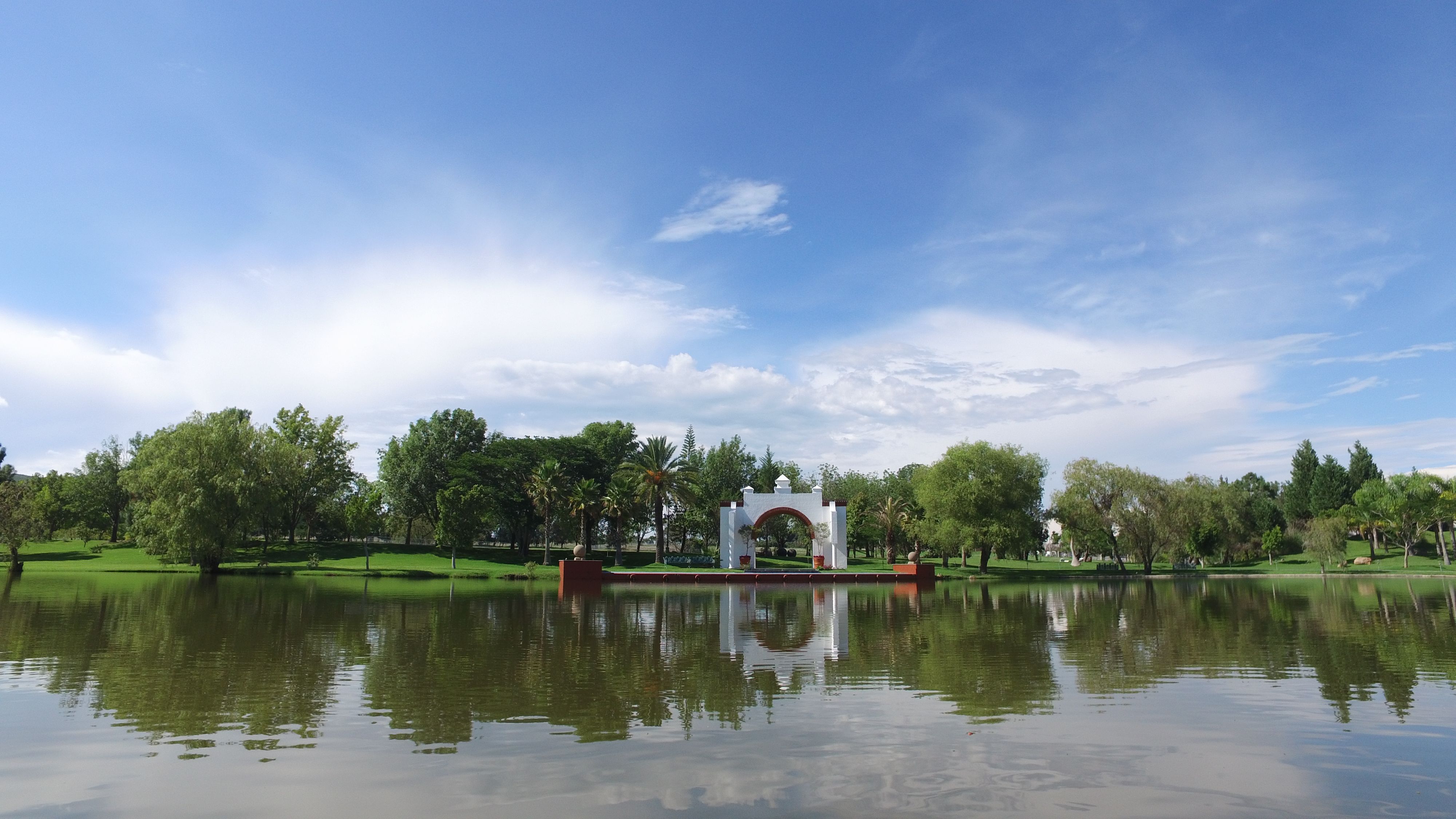 A tranquil park scene featuring a lake, trees, and a white archway reflecting in the water.
