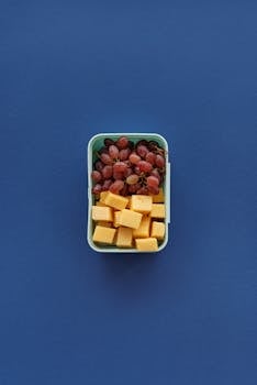 Flat lay of grapes and cheese cubes in a lunchbox on a blue background.