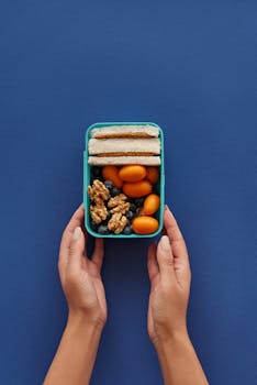 Top view of a lunchbox with sandwiches, nuts, and fruits held by hands on a blue background.