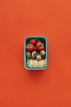 Vibrant flat lay of strawberries, quail eggs, and biscuits in a blue box on an orange background.
