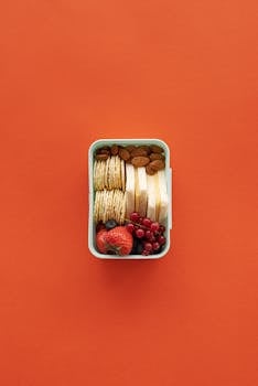Flat lay of a nutritious lunchbox with crackers, almonds, fruits on an orange background.