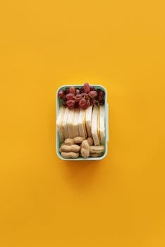 Top view of a lunch box with grapes, sandwich, and peanuts on yellow background.