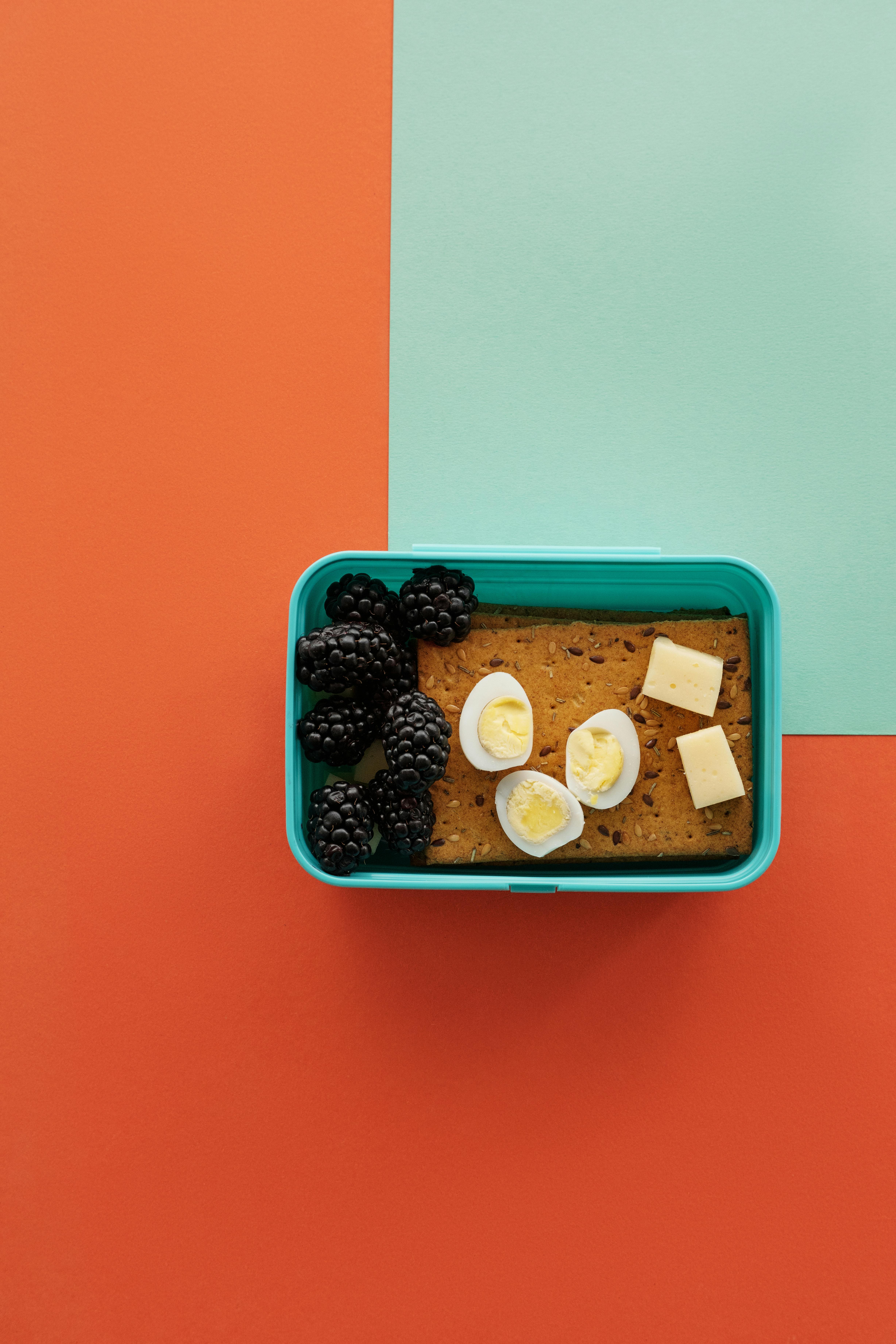 橙色背景上，摆放着五颜六色的午餐盒，里面装有黑莓、煮鸡蛋和奶酪