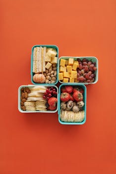 Top view of four lunch boxes with fruits, cheese, and snacks on orange background.