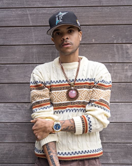 African American man wearing knitted sweater, necklace, and black cap standing against a wooden wall.