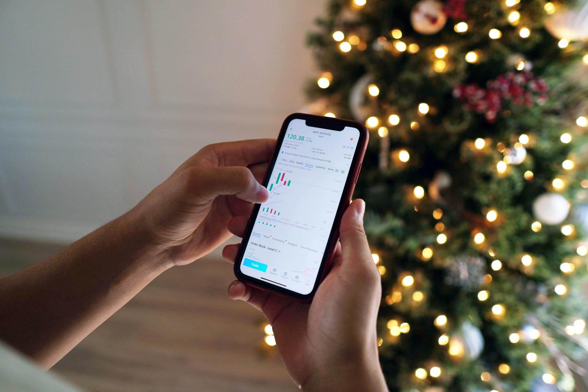 Close-up of a person using a stocks app on a smartphone in front of a decorated Christmas tree.