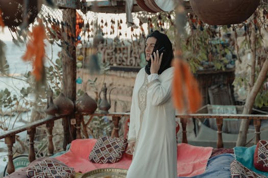 Woman wearing hijab and traditional dress, talking on phone in ornate outdoor setting.