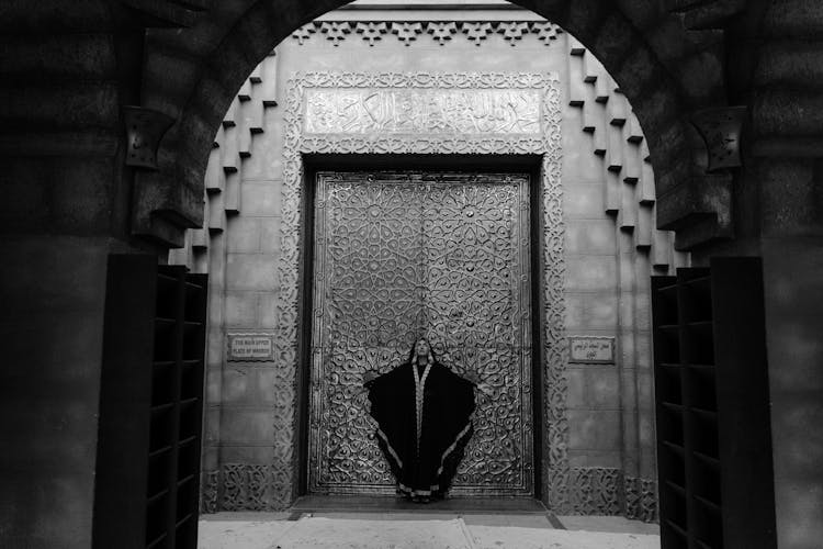 A Grayscale Of A Woman In A Robe At The Entrance Of A Building