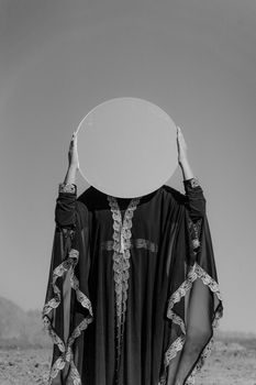 A woman in a traditional abaya holding a mirror against a clear sky, artistic and thought-provoking.