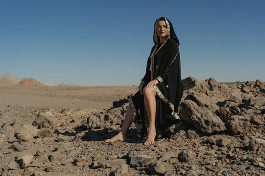 在湛蓝的天空下，一位身着传统服装的女人坐在沙漠中的岩石上